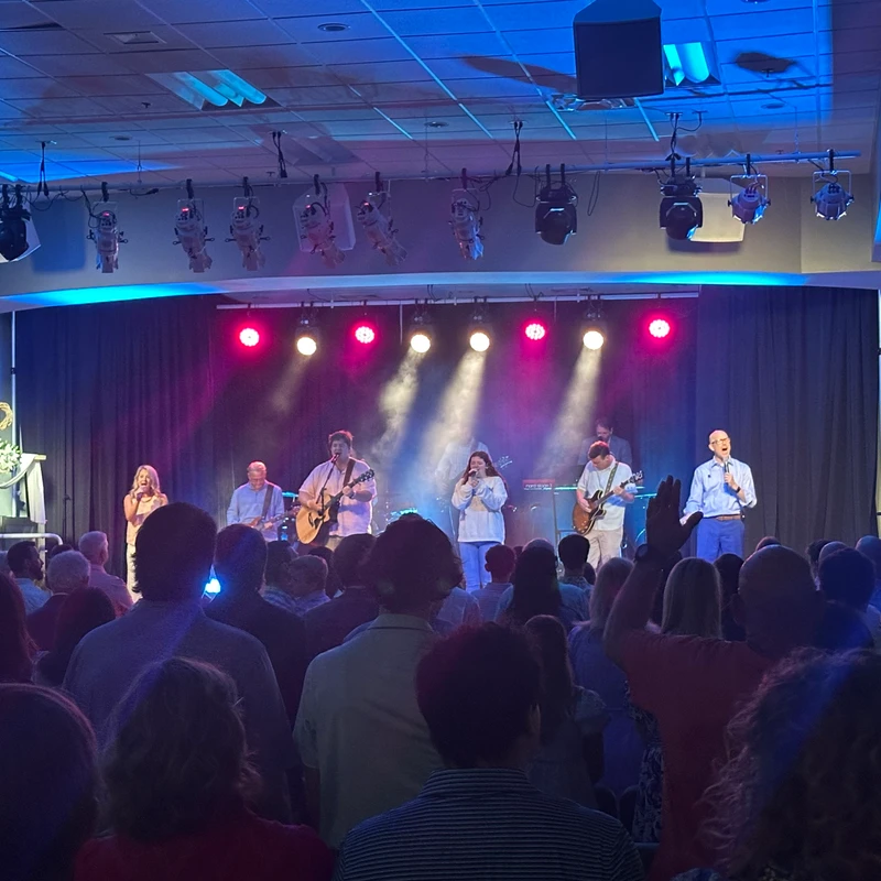 A view from the audience of a worship band performing on a brightly lit stage during a church service, with some attendees raising their hands.