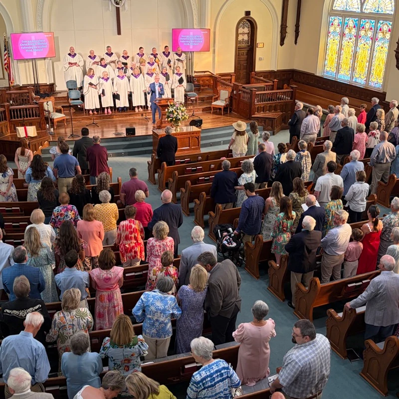 A high-angle view of a full church sanctuary. The congregation stands in wooden pews, facing a choir in robes and a speaker at the podium during a service.