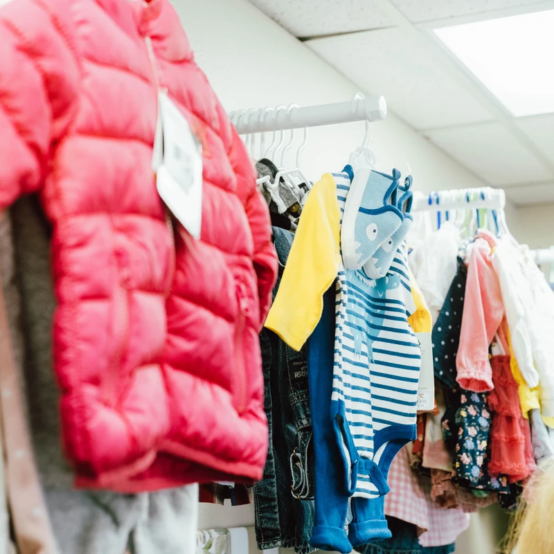 A close-up of a clothing rack filled with colorful children's clothes. A pink puffer jacket and a blue striped onesie are in the foreground.