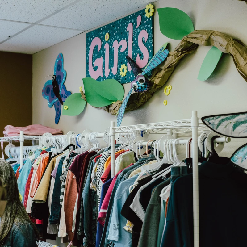 A clothing rack filled with girls' clothes sits under a colorful, decorated wall with a 'Girl's' sign and handmade paper butterflies and dragonflies.