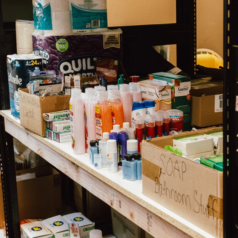 Storage shelves are filled with donated toiletries like shampoo, soap, and toothpaste, along with large packs of toilet paper, organized in cardboard boxes.
