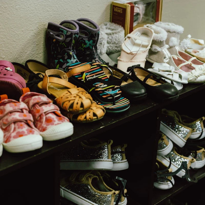Many pairs of colorful children's shoes, including sneakers, boots, and sandals, are neatly arranged on two levels of a dark-colored shelving unit.