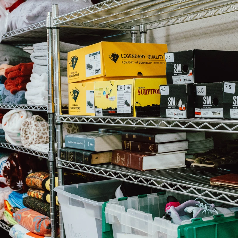 Metal wire shelves are filled with donated items including folded blankets, shoeboxes, books, and bins of clothing in a storage room.
