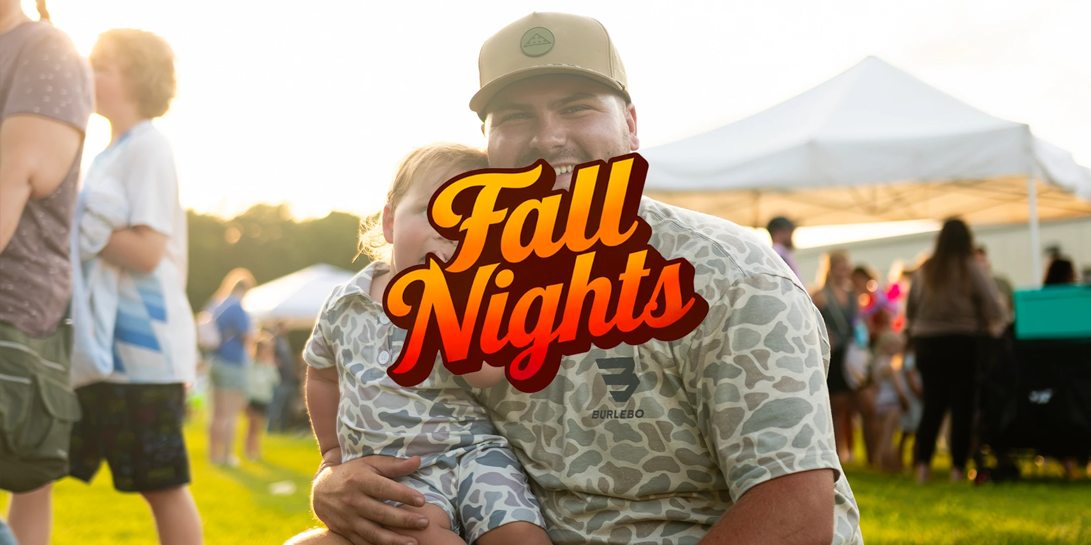 A smiling man holds a young child on his lap at an outdoor event at sunset. They are both wearing camouflage. The text 'Fall Nights' is over the image.