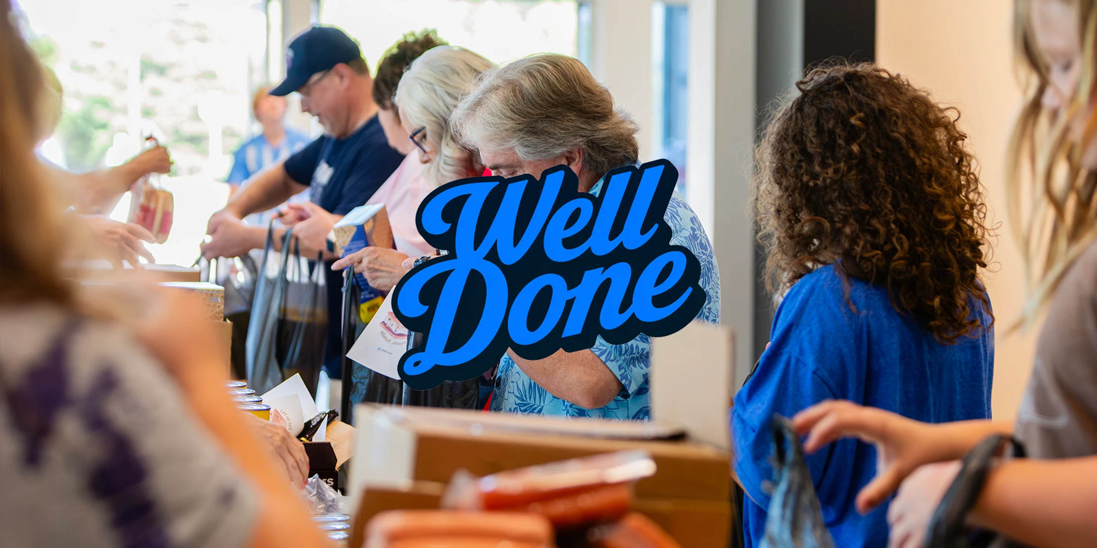 A group of volunteers stand in a line at a long table, packing donated items into bags. A large blue 'Well Done' graphic is overlaid on the image.