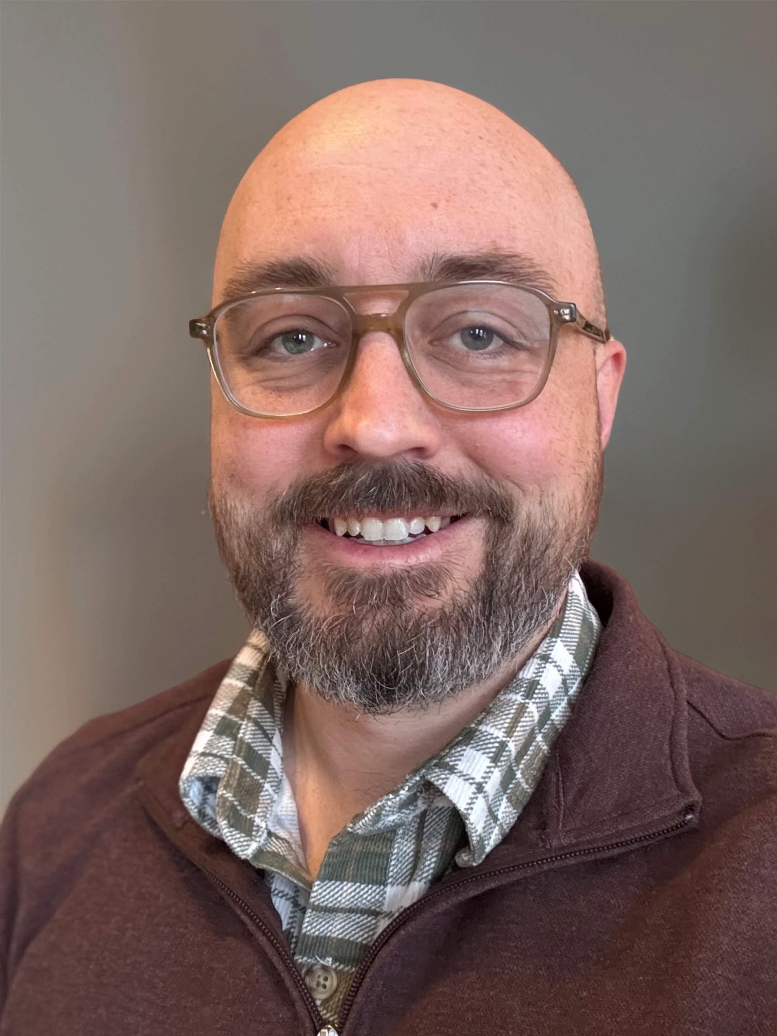 A headshot of a bald man with a salt-and-pepper beard and glasses. He is smiling and wearing a plaid shirt under a brown zip-up sweater.