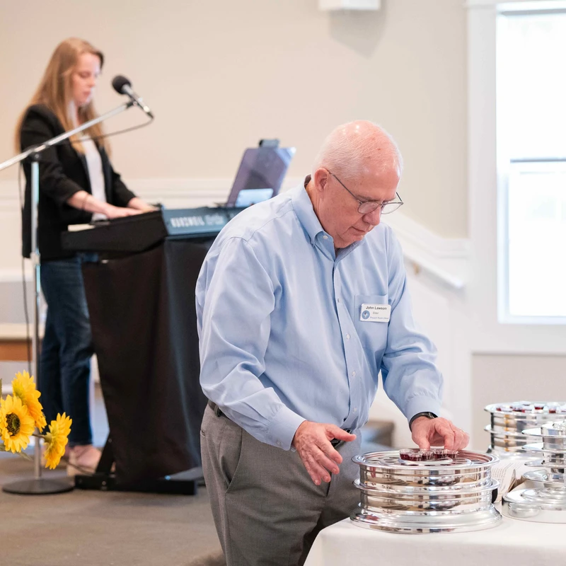 An older man in a blue shirt and glasses arranges small cups on a silver communion tray, with a woman playing a keyboard in the background.