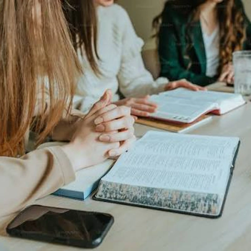 Three women sit around a wooden table with open books, appearing to be in a Bible study or prayer group. One woman has her hands clasped in front of her.