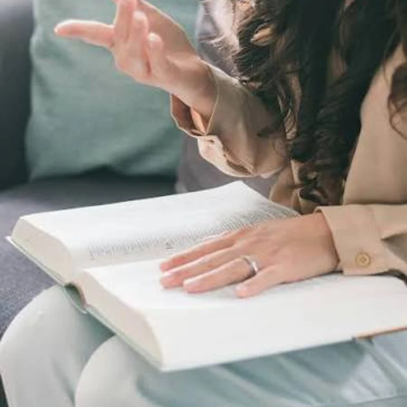 A woman sits on a couch with an open book on her lap. One hand is on the page, and the other is gesturing as if she's talking about what she's reading.