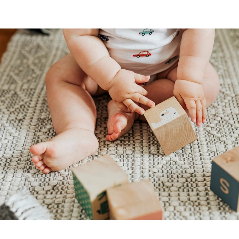 A close-up of a baby in a car-patterned onesie sitting on a textured rug and playing with wooden blocks with letters and pictures on them.