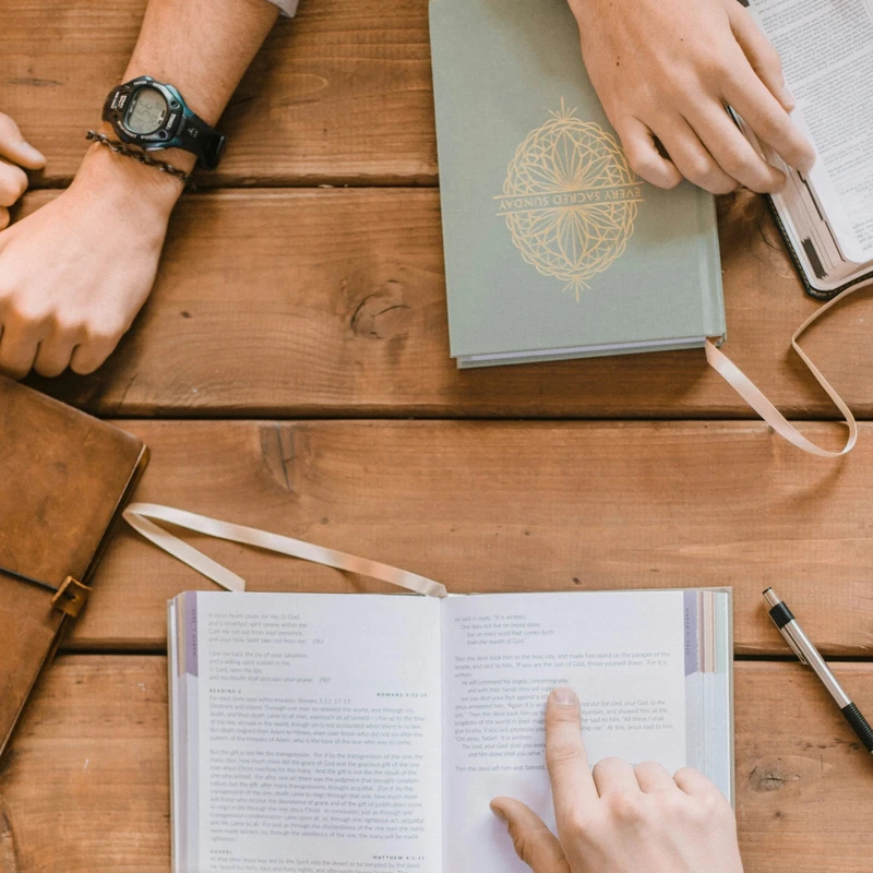 Overhead view of two people studying together at a wooden table. One person points at a passage in an open book, while other books rest nearby.