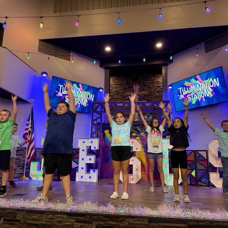 A group of children stand on a brightly decorated stage with their hands in the air, participating in a church event called 'Illumination Station'.