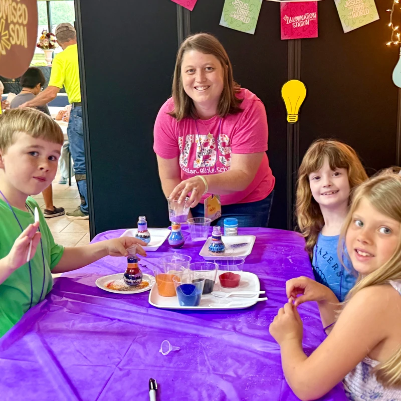 An adult woman helps three smiling children at a table make colorful sand art in bottles during a craft activity at what appears to be a church event.