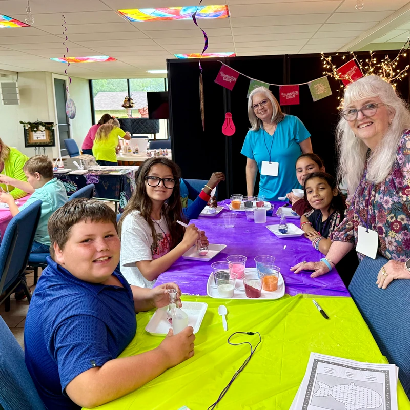 A group of smiling children and adults gathered around a table with colorful tablecloths, participating in a hands-on craft activity in a decorated room.