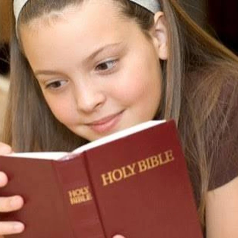 A young girl with long brown hair and a white headband lies down while intently reading from a red book titled 'Holy Bible'.