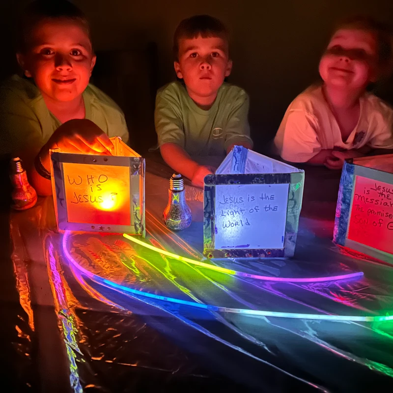 Three children in a dark room illuminated by homemade lanterns with handwritten messages about Jesus. Colorful glow sticks are on the table in front of them.