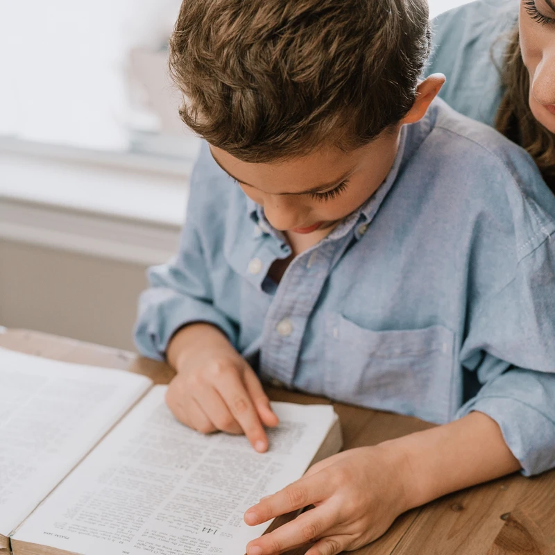 A young boy in a blue shirt points to text in a large open book while a woman, likely his mother, looks over his shoulder at a wooden table.