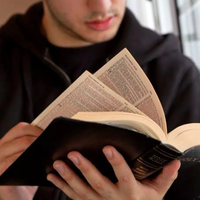 A close-up of a young man in a black hoodie holding an open Holy Bible. The focus is on the book and his hands, with pages from the Gospel of Matthew visible.