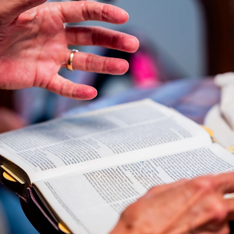 A close-up shot of a person's hands holding an open Bible. One hand is raised in a gesture over the text, with a gold ring visible on the finger.