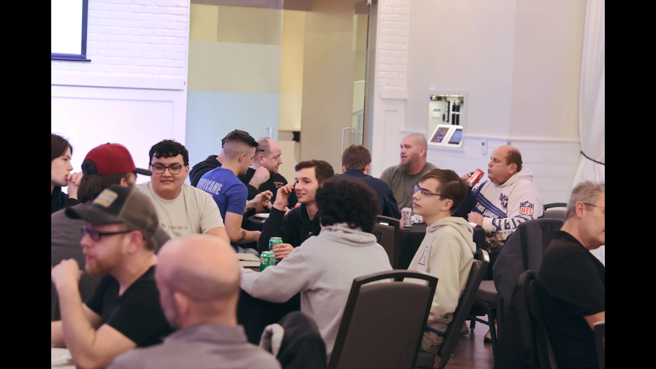 A group of casually dressed men of various ages sit at round tables in a well-lit room, talking and interacting with each other during a gathering.