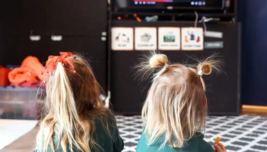 The backs of two young blonde girls sitting on a patterned rug watching TV. One has a red bow in her hair, and the other has two small pigtails.