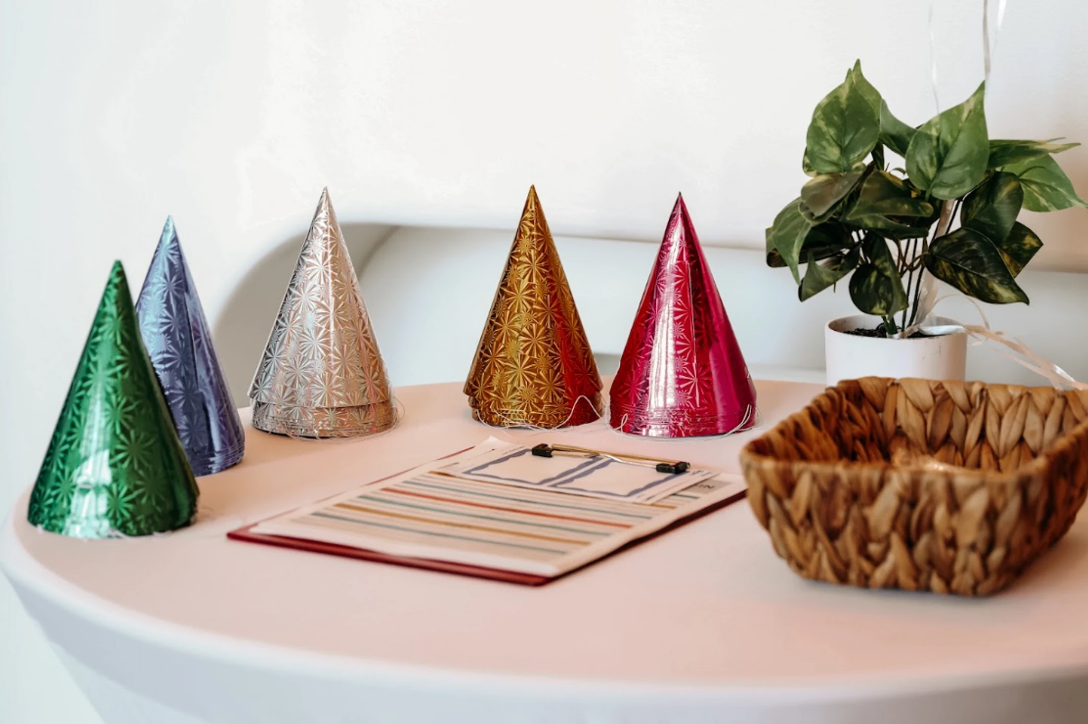 Five shiny party hats in green, blue, silver, gold, and red sit on a white table next to a clipboard, a woven basket, and a small potted plant.