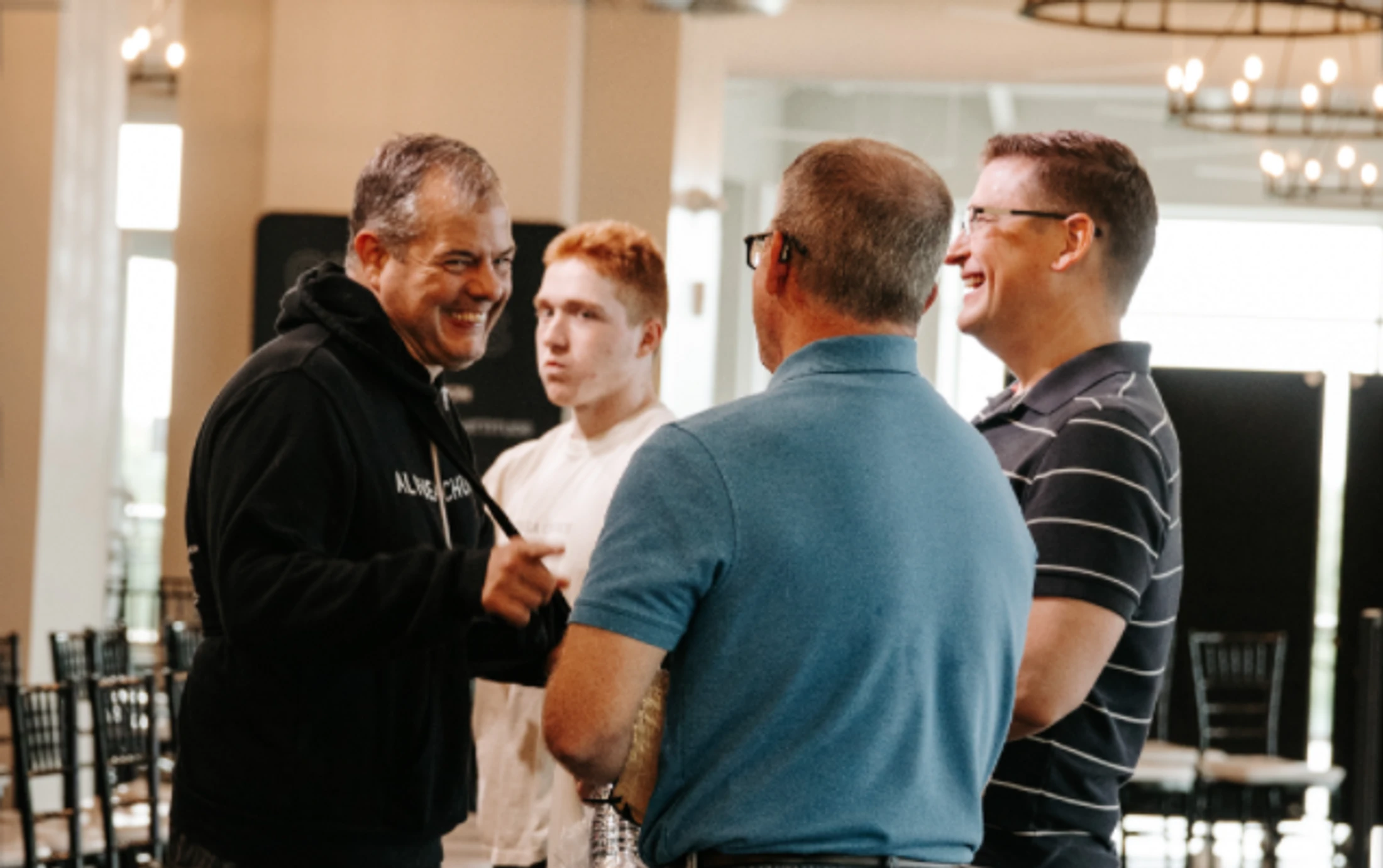 Four men stand in a circle talking at an indoor event. The man in the black hoodie on the left is smiling widely, and the man on the right is also smiling.