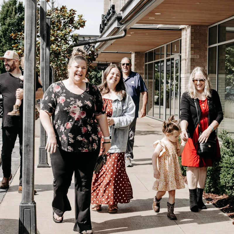 A multi-generational family smiles and laughs while walking together on a sunny city sidewalk in front of a building.