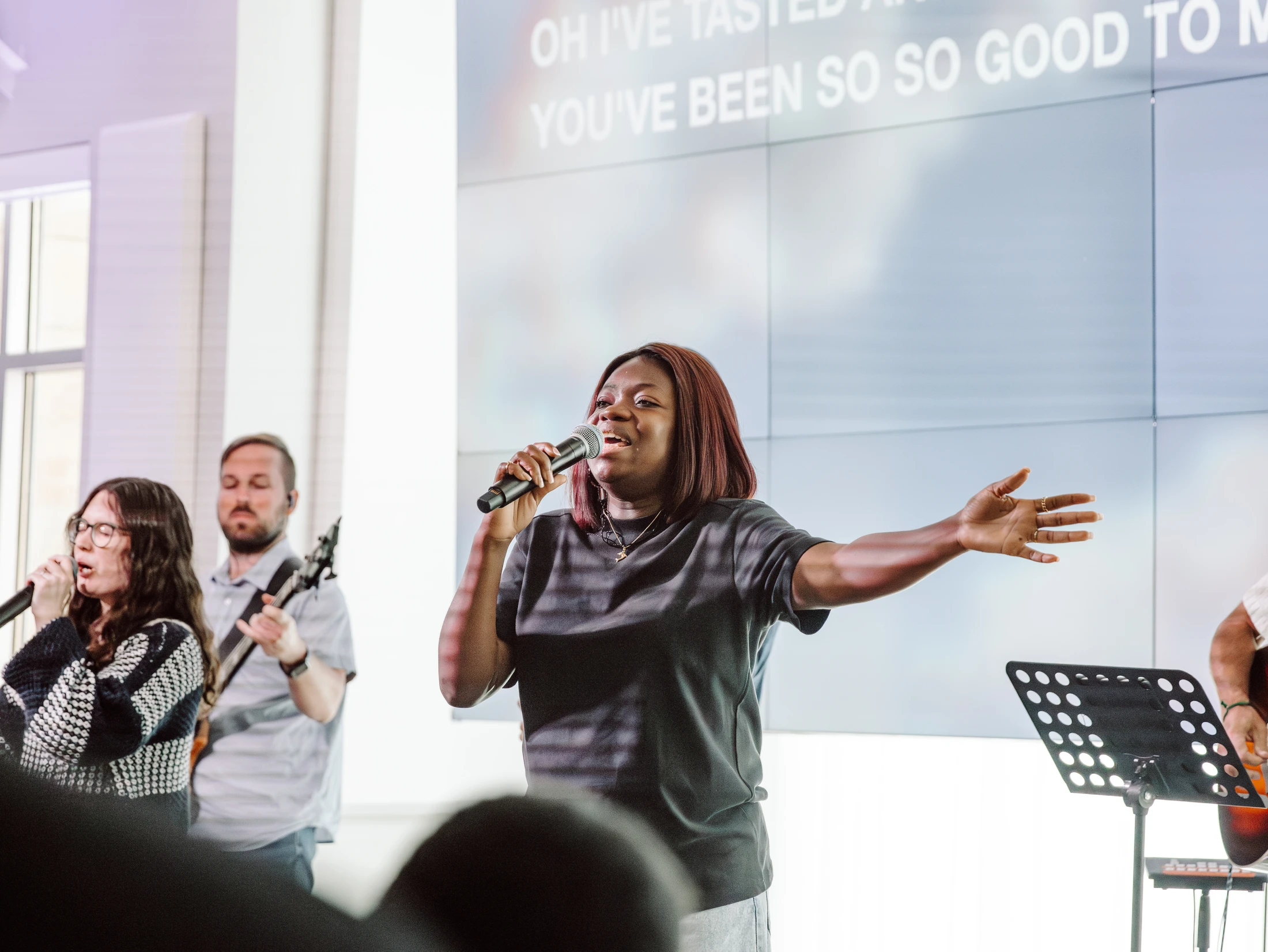 A Black woman sings into a microphone with her arm outstretched, leading a band on a brightly lit stage with lyrics projected on a screen behind her.