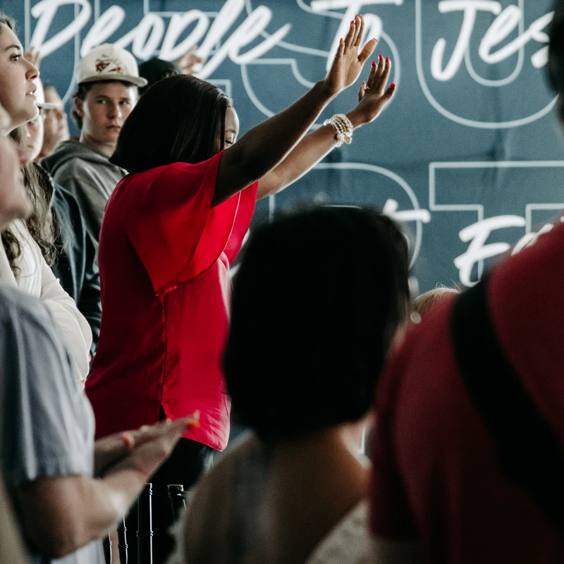A woman in a red shirt stands in a congregation, her hands raised in worship. Behind her is a blue wall with the words 'People to Jesus' in white.