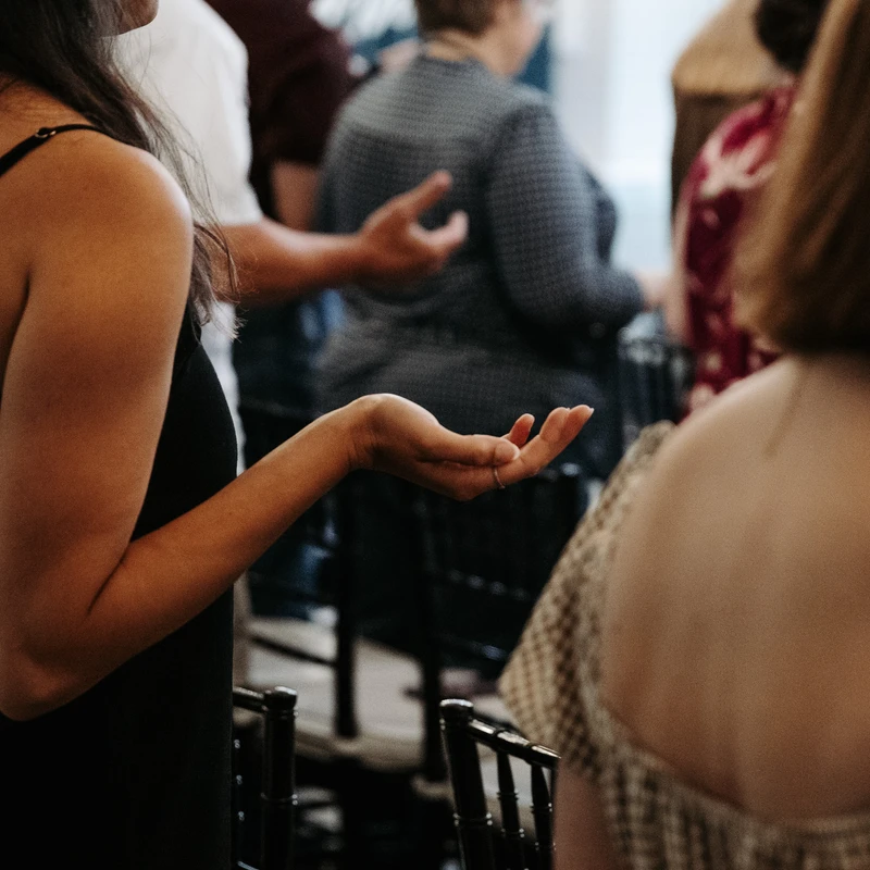 A side view of a woman in a black top with her hand outstretched, palm up, in a gesture of worship or prayer during a gathering with other people.