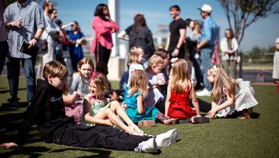 A group of children sit together on a green lawn while adults stand and socialize in the background on a sunny day.