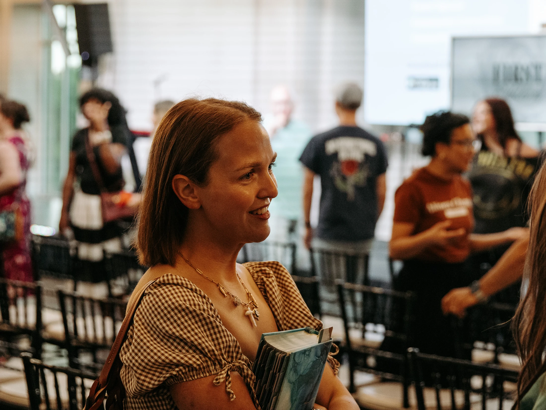 A woman with light brown hair smiles while holding a book at an indoor gathering. She is wearing a gingham top and a necklace, with people blurred behind her.