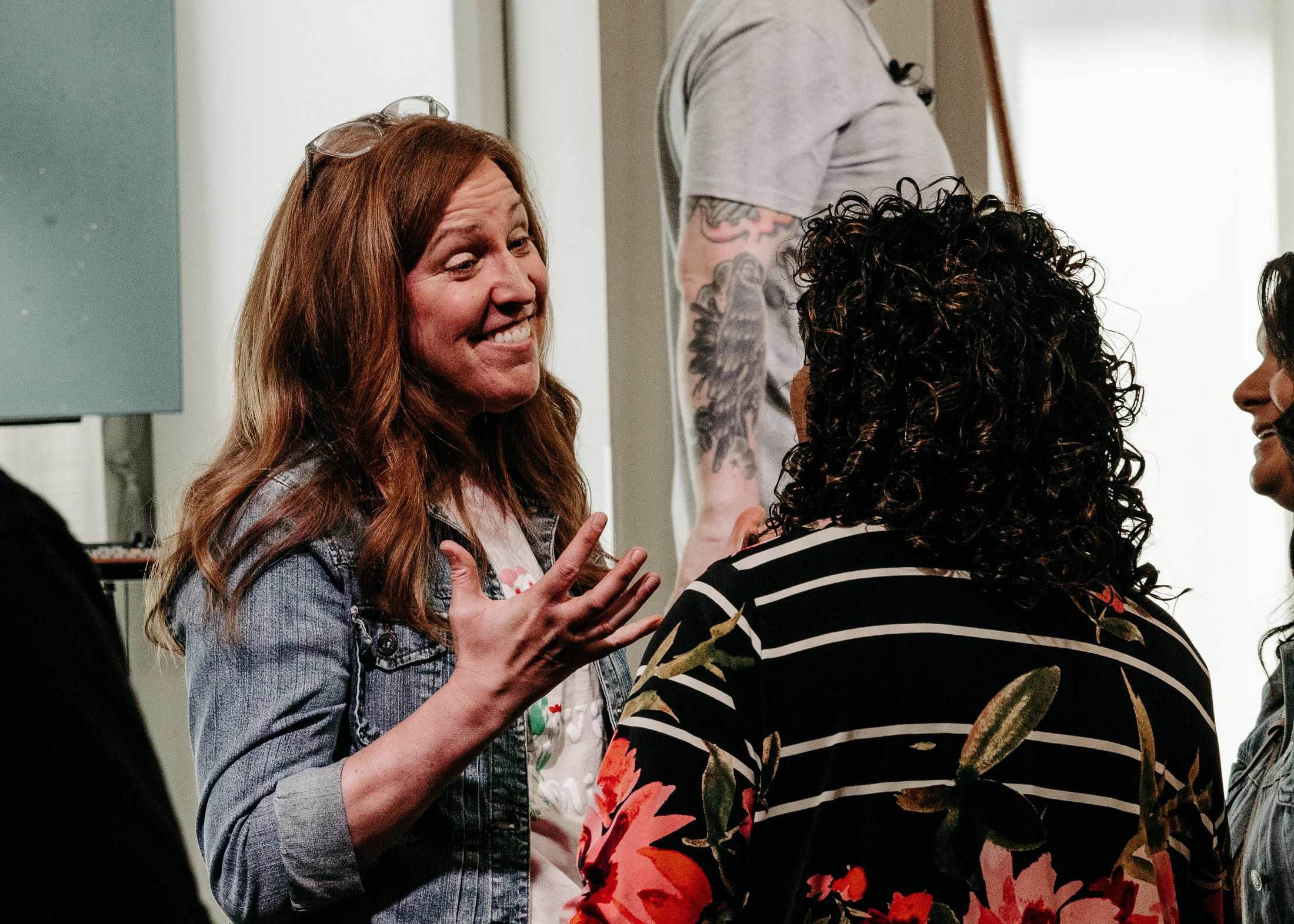 A smiling woman with long reddish-brown hair and a denim jacket gestures with her hands as she speaks to another woman with curly hair, who is seen from behind.