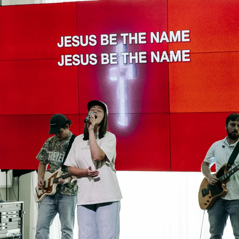 A female singer and a band perform on stage in front of a large red screen with the words 'Jesus Be The Name' and a glowing cross.