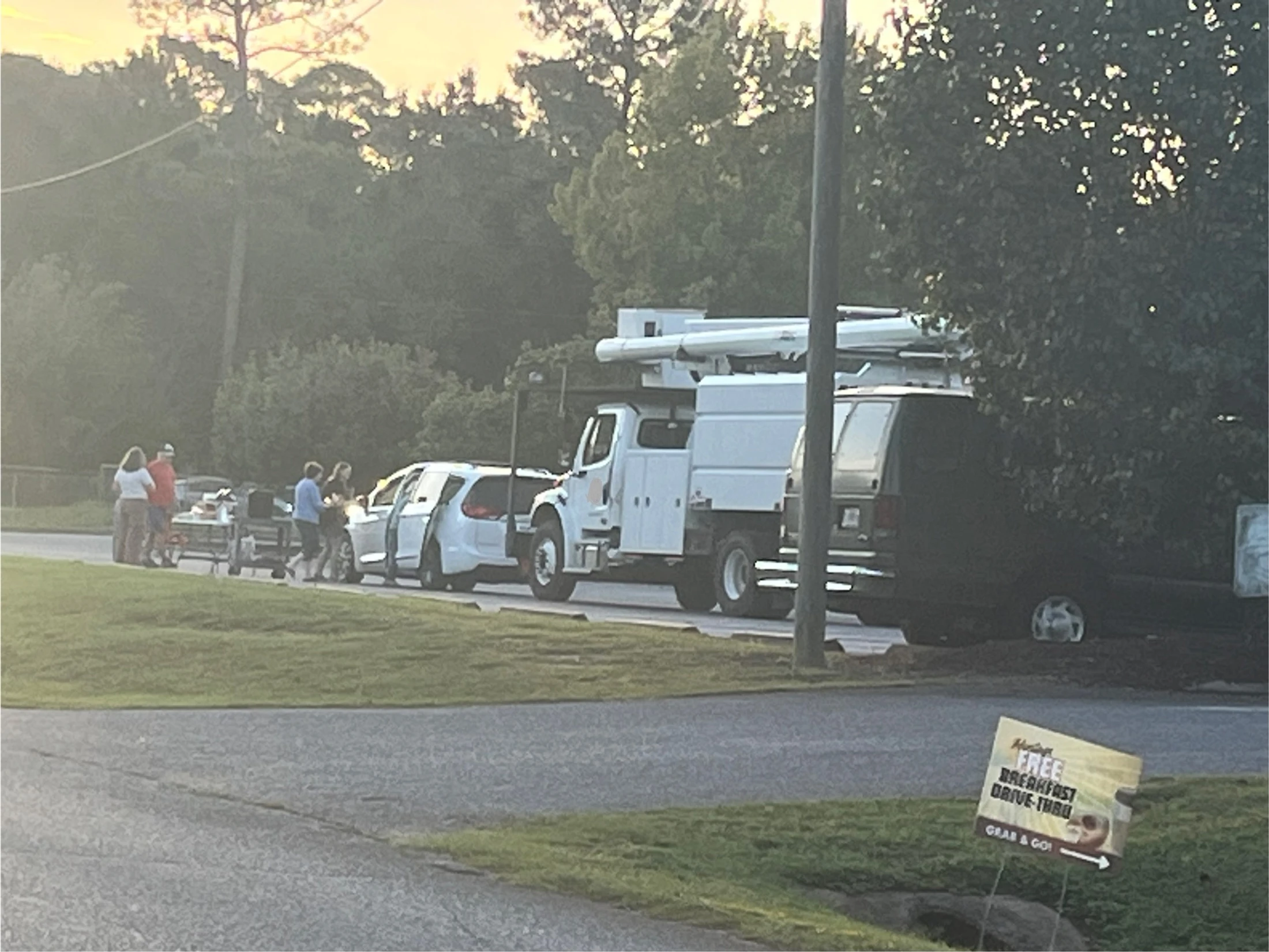 A group of people gathered for a free breakfast drive-thru event on a roadside, with a utility truck and cars parked nearby and a sign indicating the giveaway.