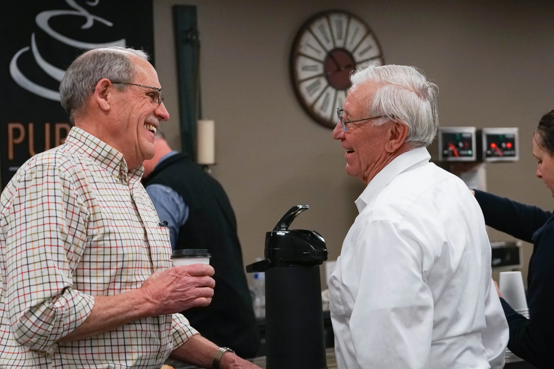 Two older men with glasses are smiling and talking to each other indoors. The man on the left wears a plaid shirt and holds a coffee cup.