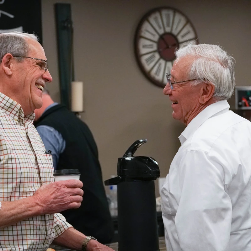 Two older men with glasses are smiling and talking to each other indoors. The man on the left wears a plaid shirt and holds a coffee cup.