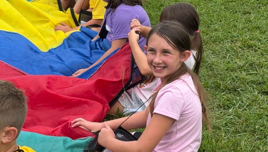 A group of children sit in a row on the grass, holding the edge of a large, colorful parachute. A girl in a pink shirt smiles at the camera.