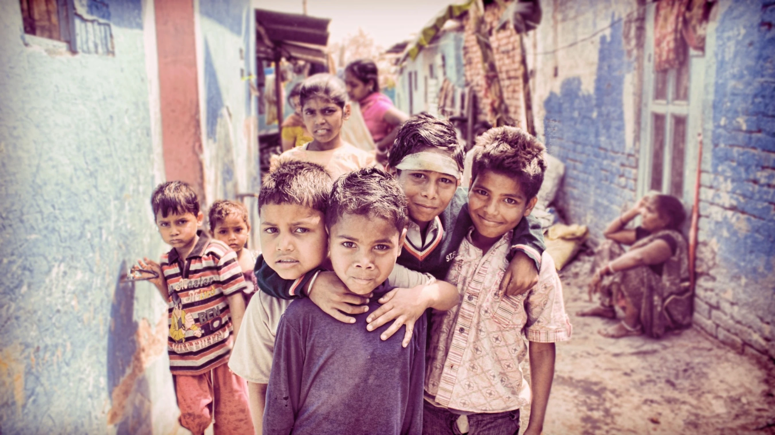 A group of young children huddle together and look at the camera while standing in a narrow alley with weathered blue and red walls.