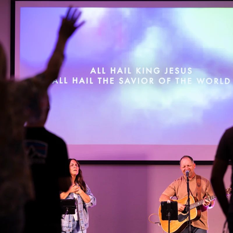 A band leads a church service with a man on guitar and a woman singing. Behind them, a screen shows lyrics. Silhouettes of the audience watch and worship.