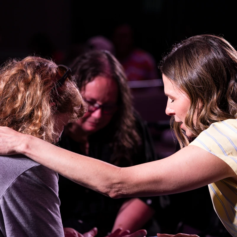 A woman with brown hair comforts another woman with curly hair by placing a hand on her shoulder as they bow their heads together in a dimly lit room.