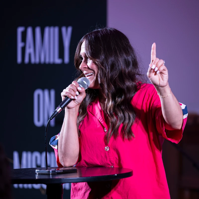 A smiling woman in a bright pink shirt speaks into a microphone on stage at Sozo Church, gesturing with her index finger pointed upward.