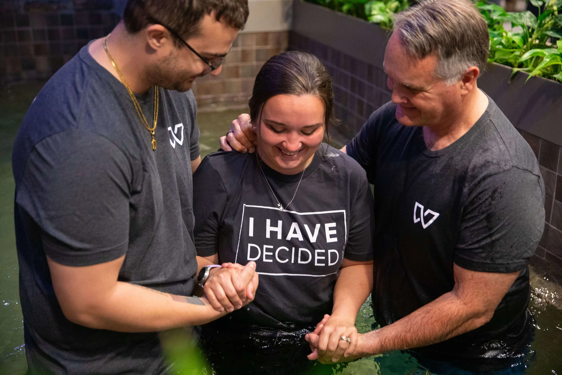 A smiling woman wearing a t-shirt that says 'I HAVE DECIDED' is supported by two men as she is baptized in a pool of water during a religious ceremony.