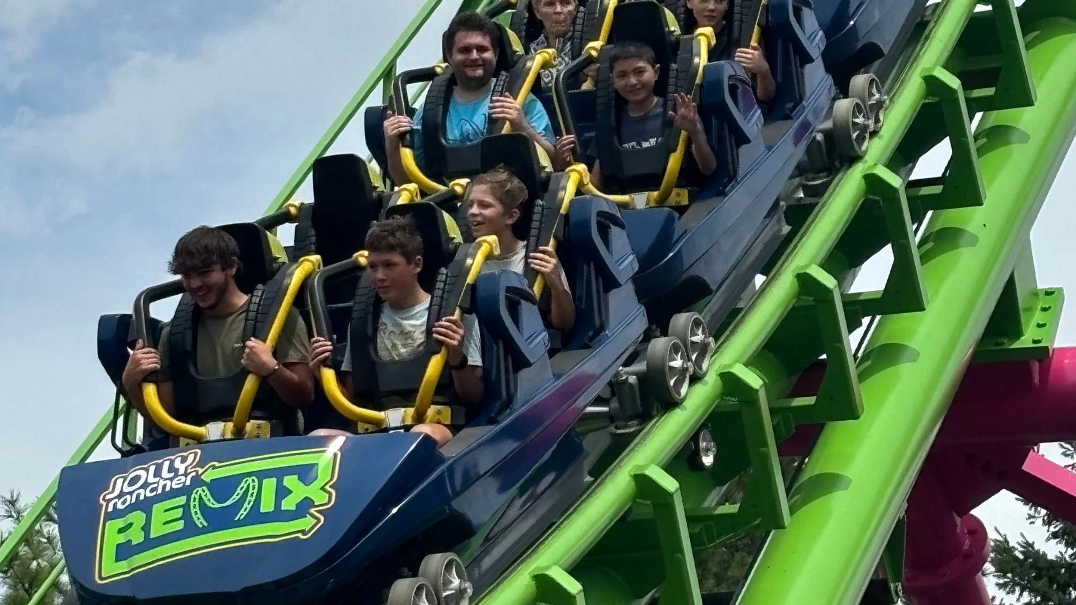 A group of smiling people riding the blue Jolly Rancher Remix roller coaster as it climbs a steep, bright green hill under a partly cloudy sky.