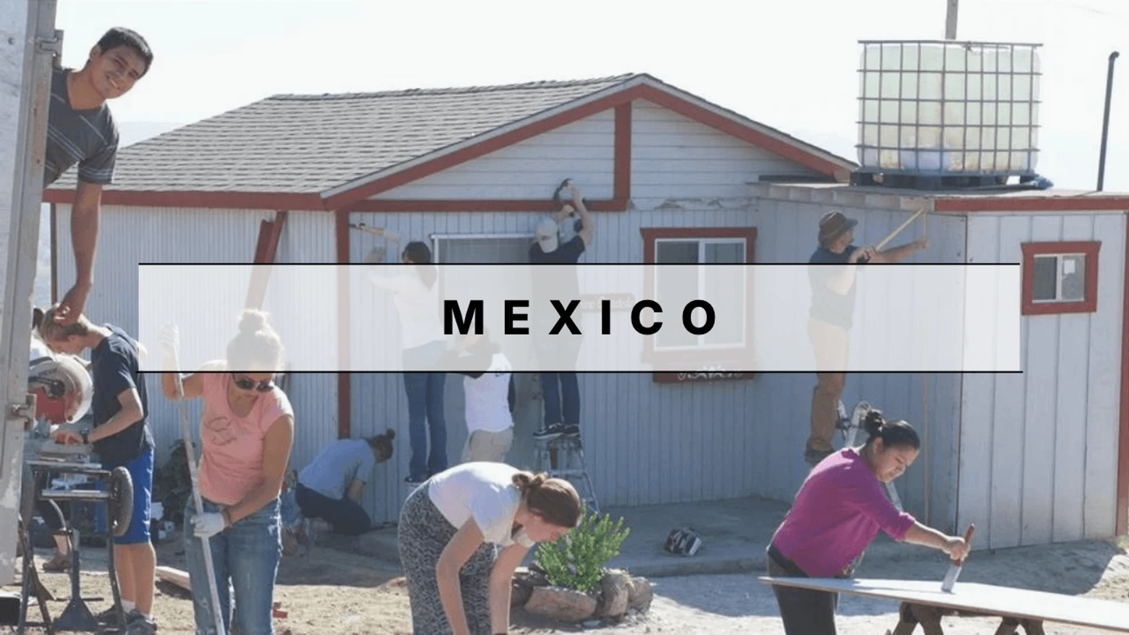 A group of young volunteers works together on the exterior of a building in Mexico, painting, sawing, and making repairs on a sunny day.