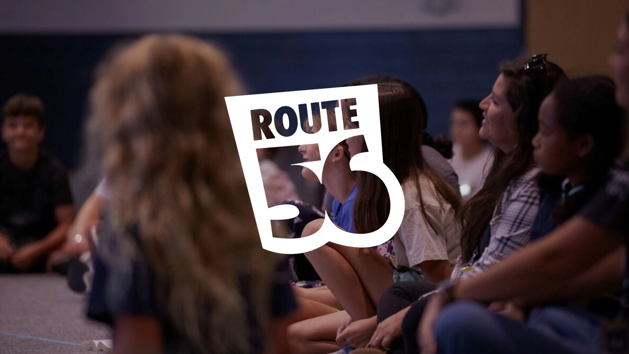 A group of young students are sitting on the floor listening during a meeting, with a white 'Route 56' logo overlaid in the center of the image.