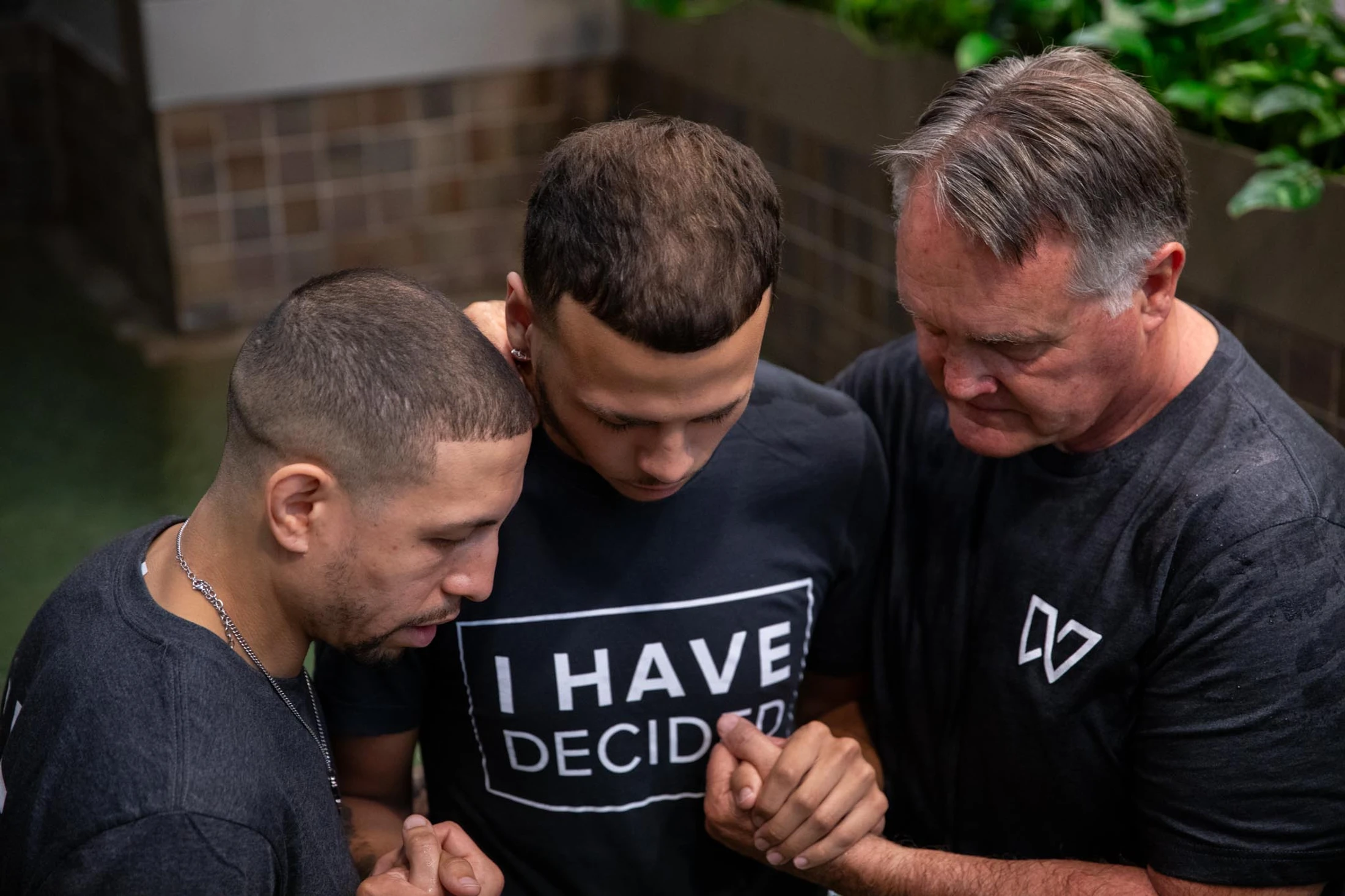 High-angle shot of three men with heads bowed in prayer. The man in the center wears a shirt that reads 'I HAVE DECIDED' as two others support him.