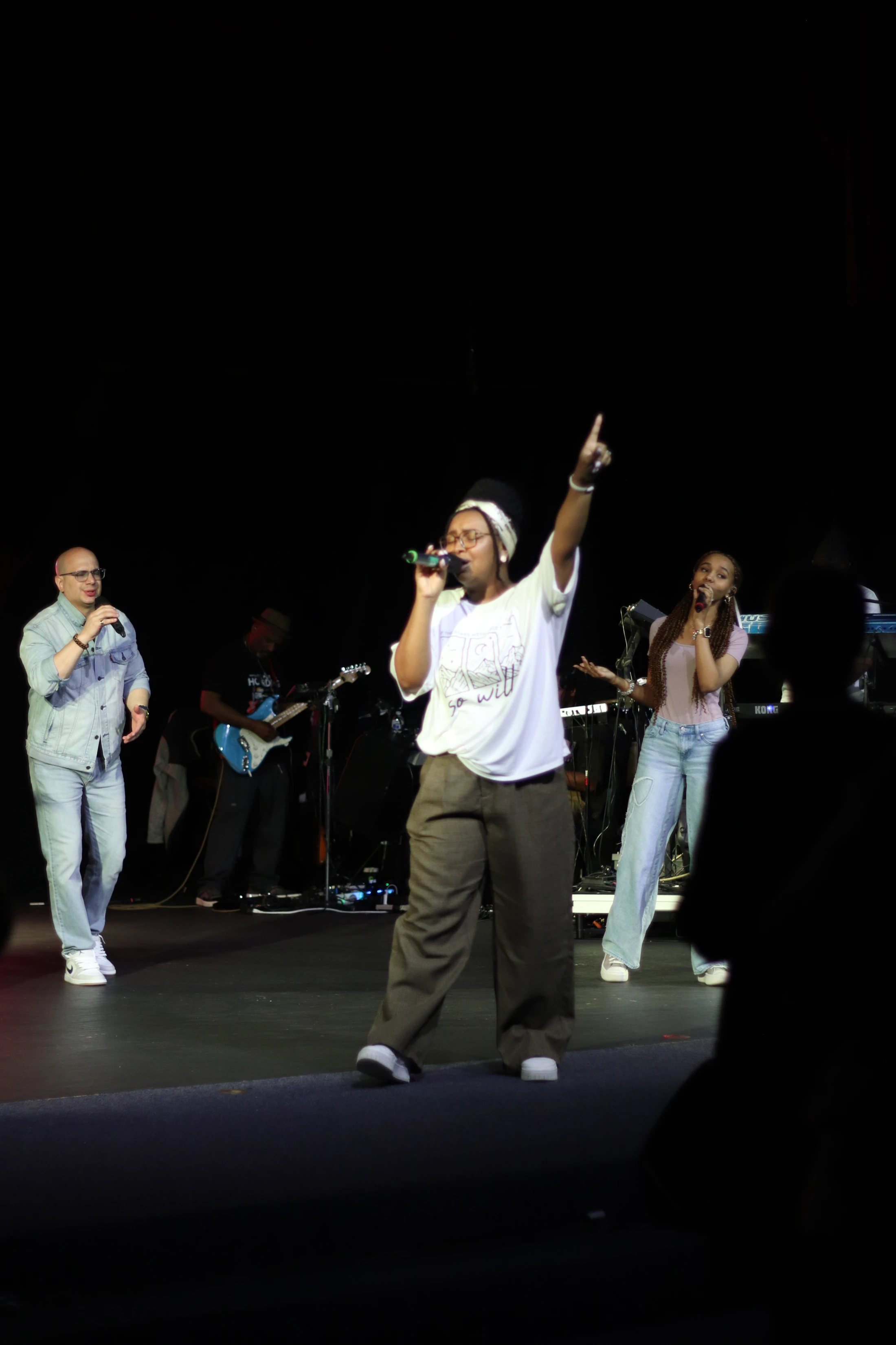 A band with three singers and a guitarist performs on a dimly lit stage. The lead singer in the center holds a microphone and points towards the ceiling.