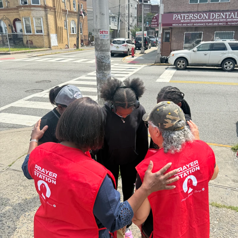 A group of people prays in a circle on a city sidewalk. Two volunteers in red "Prayer Station" vests place supportive hands on the others, who bow their heads.